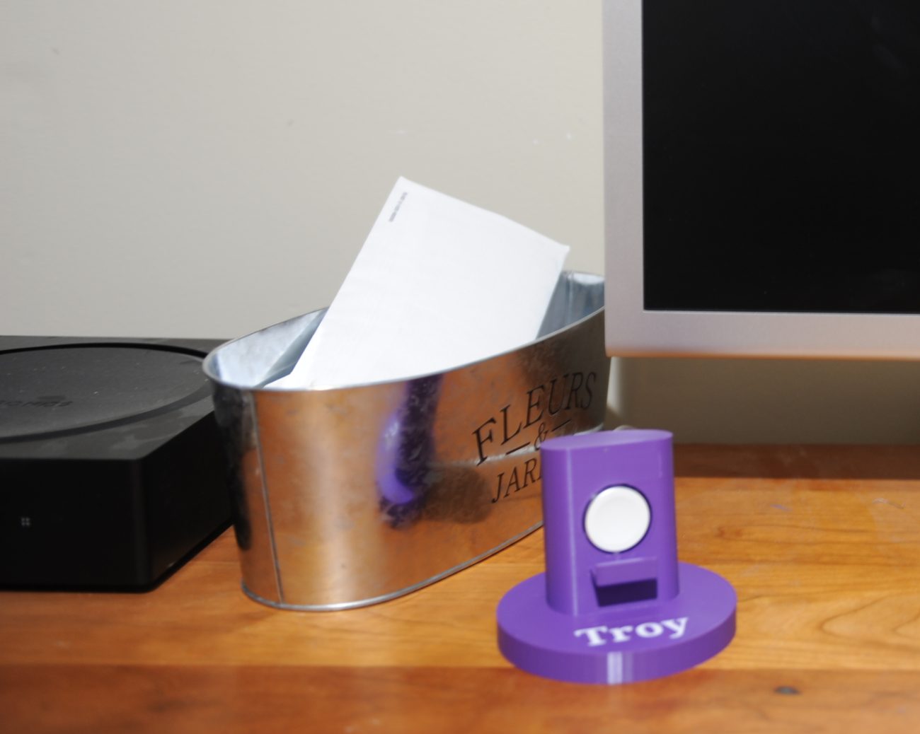 A purple Apple Watch stand with the name Troy on it, on a wooden desk next to a tin bucket