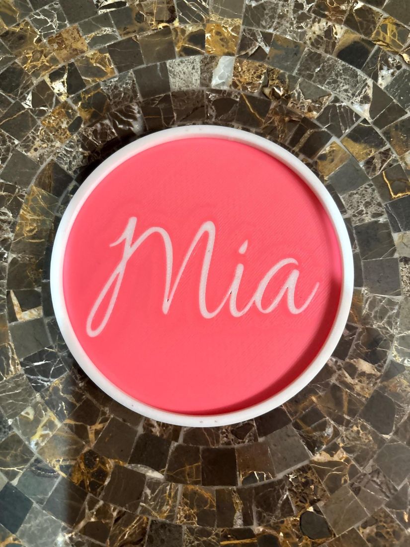 A round coral coaster with a name in white script